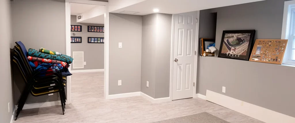 Gray finished basement interior in Marlboro MA showing recessed lighting, stacked chairs, framed sports artwork, storage door, and clean open floor space.