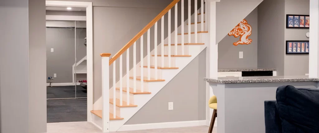 Modern finished basement in Marlboro MA with gray walls, wooden staircase with white railing, recessed lighting, and granite countertop bar seating.