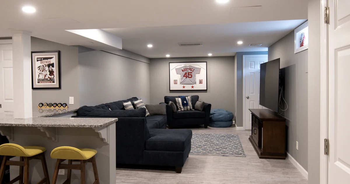 Finished basement living area in Marlboro MA featuring gray walls, recessed lighting, navy sectional sofa, granite countertop bar, and wall mounted TV.