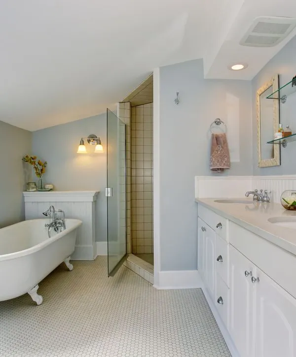A traditionally renovated bathroom by remodeling contractors in Auburn, MA, featuring a white clawfoot tub, light blue walls, and a classic white vanity with penny tile flooring.