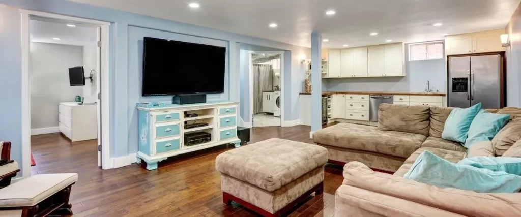 A comfortable basement family living area with light blue walls, featuring a large brown sectional sofa, a flat-screen TV on a blue stand, and a background kitchenette with a stainless steel refrigerator.