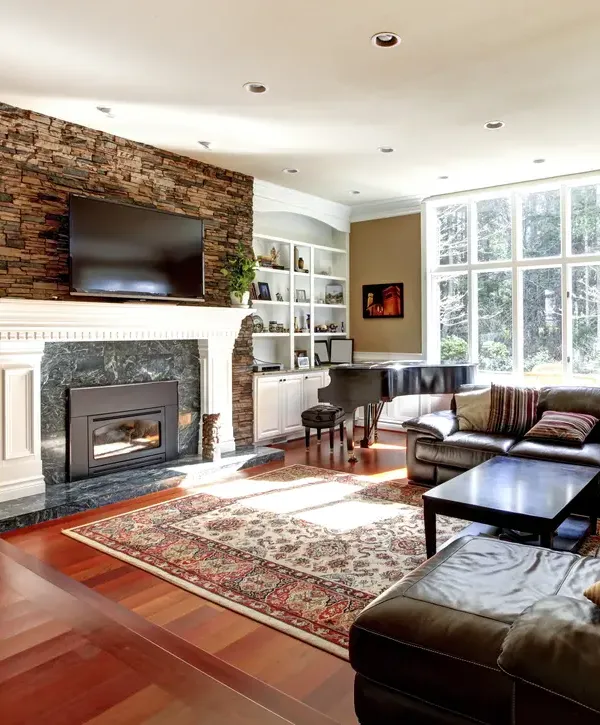 A spacious, remodeled living room featuring a custom stone fireplace, built-in white bookshelves, hardwood floors, and a grand piano, completed by design-build remodeling companies in Worcester, MA.