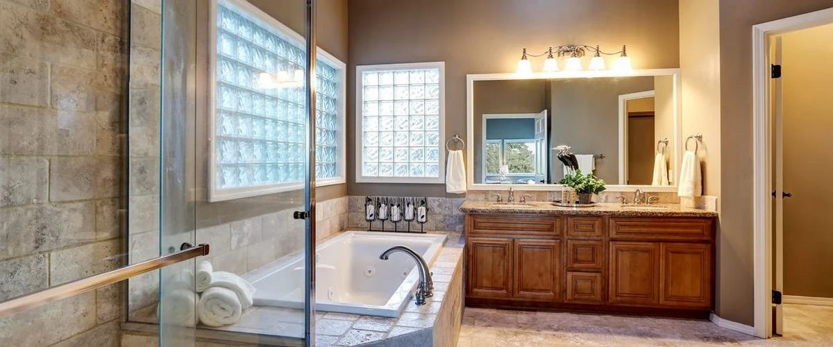 Spacious warm-toned bathroom featuring a relaxing corner soaking tub, classic glass block windows, and a large custom double vanity with rich wood cabinetry.