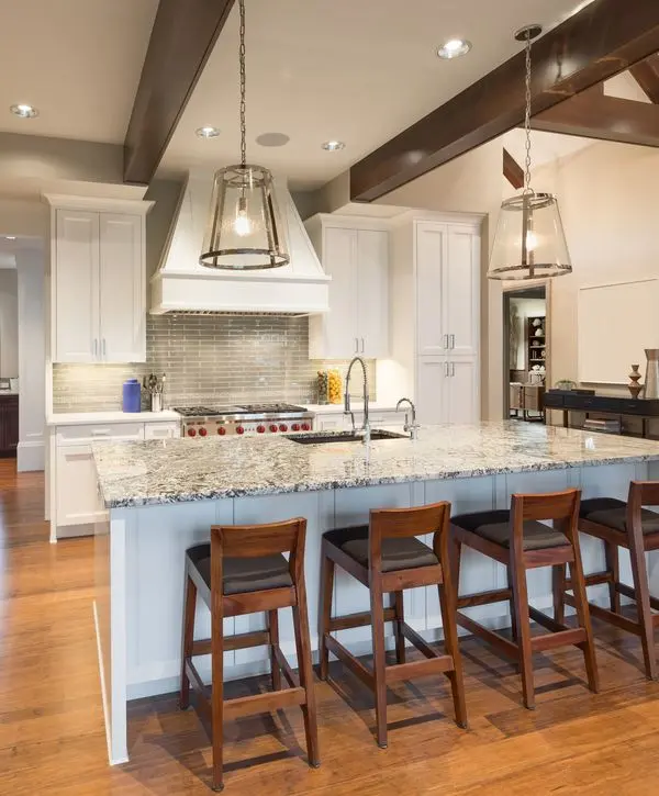 Large traditional white kitchen featuring a prominent granite island, wooden bar stools, glass pendant lights, and dark exposed ceiling beams, designed by expert kitchen remodeling contractors in Worcester, MA.
