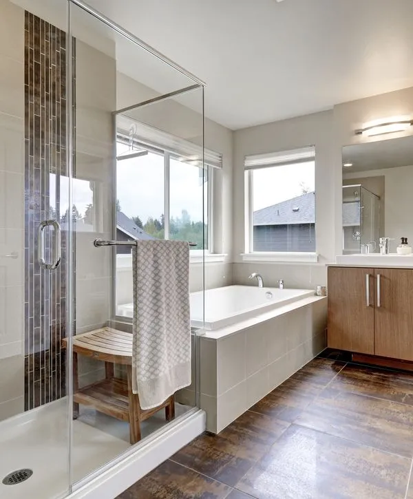 Modern bathroom featuring a glass walk-in shower with a wooden bench, a bright bathtub by the window, and a sleek vanity, finished by expert bathroom remodeling contractors in Worcester, MA.