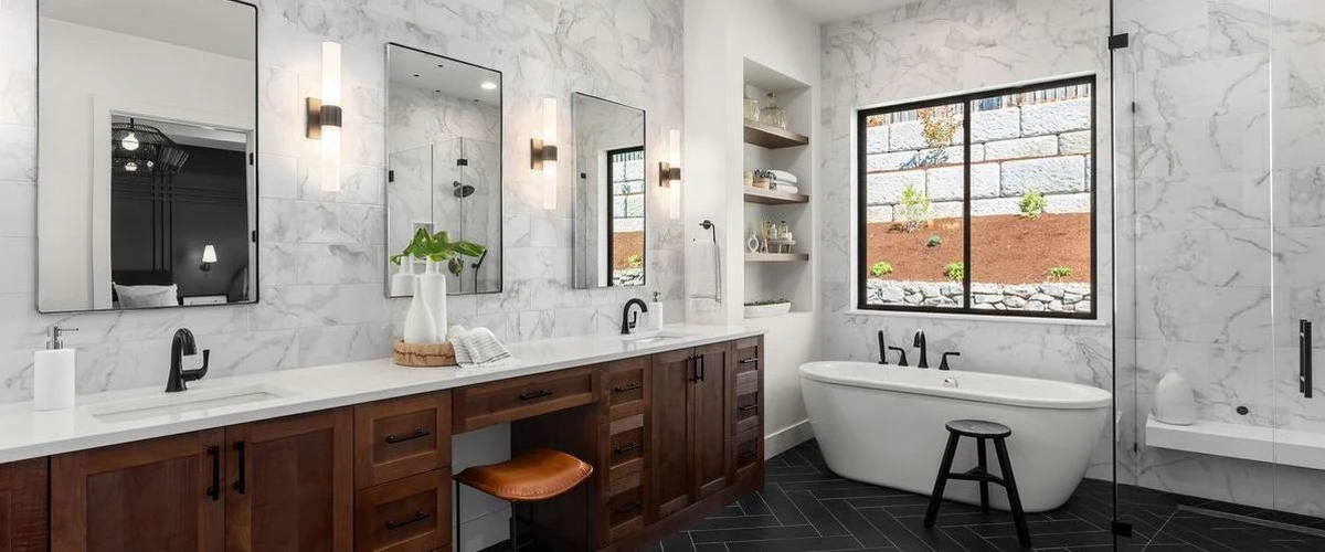A luxury modern bathroom featuring a double dark wood vanity, white marble wall tiles, black fixtures, and a freestanding tub against a black herringbone tile floor.