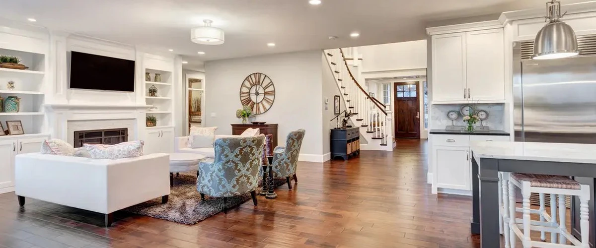 A spacious open-concept home interior featuring a white fireplace with built-in shelving, hardwood floors, and a modern kitchen with white cabinetry and a dark island.