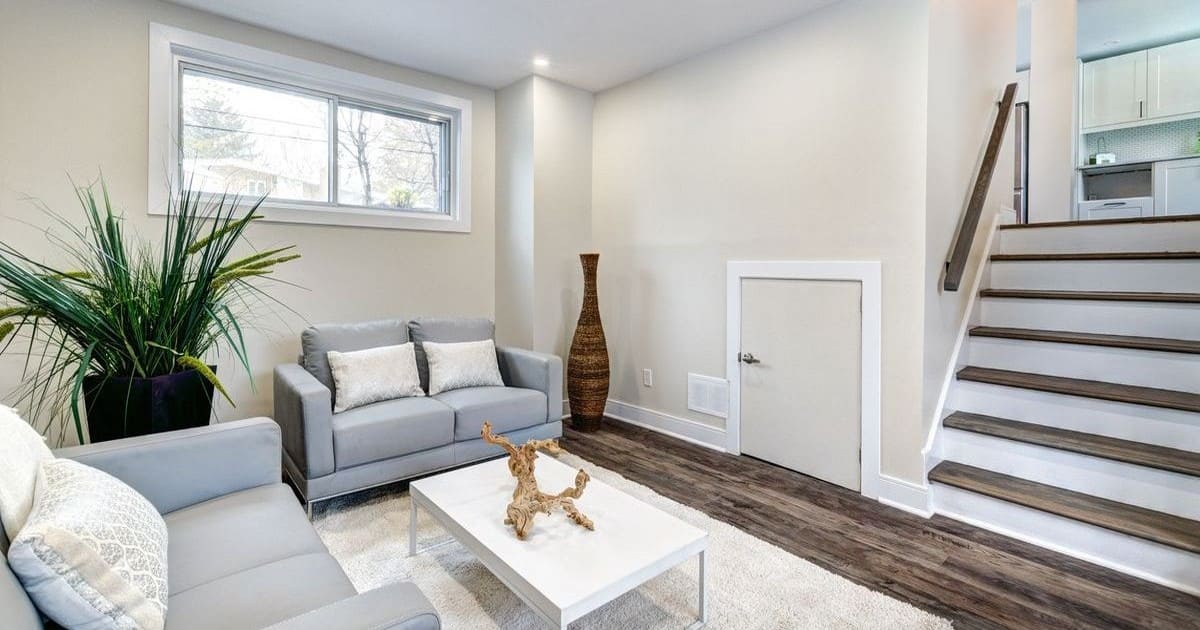 A brightly lit, modern sitting area at the bottom of a wooden staircase featuring two grey loveseats, a white coffee table, and wood-style flooring, showcasing finishes that factor into your basement remodel cost.