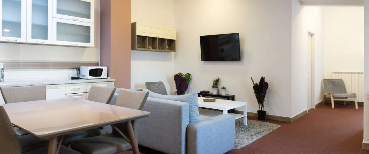 A modern basement living space featuring an open-concept layout with a small white kitchenette, grey sofa, dining table, and wall-mounted TV.