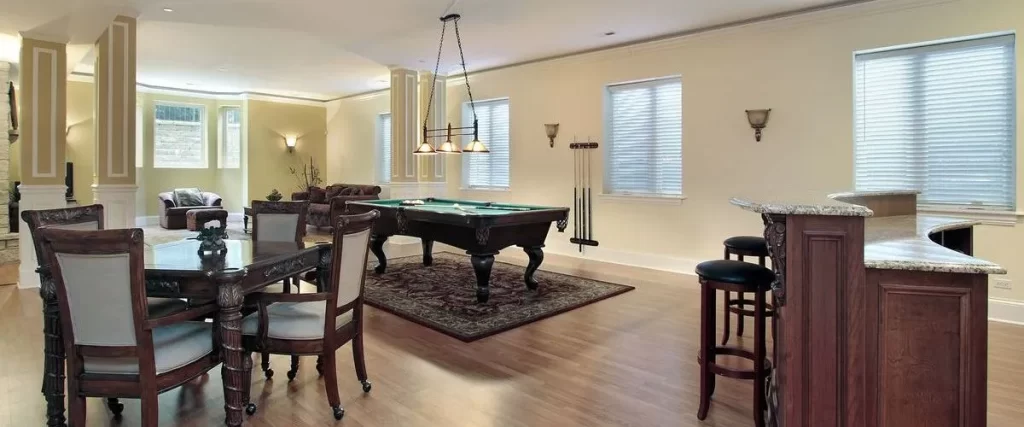 A spacious, finished basement entertainment room featuring a green felt pool table, a dark wood dining table with chairs, a wet bar area, and hardwood floors under recessed lighting.