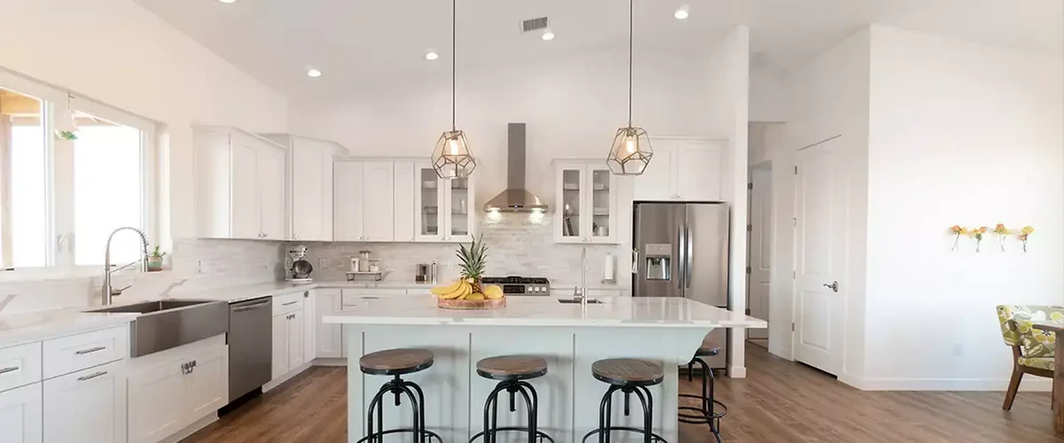 A spacious modern kitchen showing white shaker cabinets, an apron front sink, a large island with three industrial-style stools, geometric pendant lights, and a fresh fruit bowl.