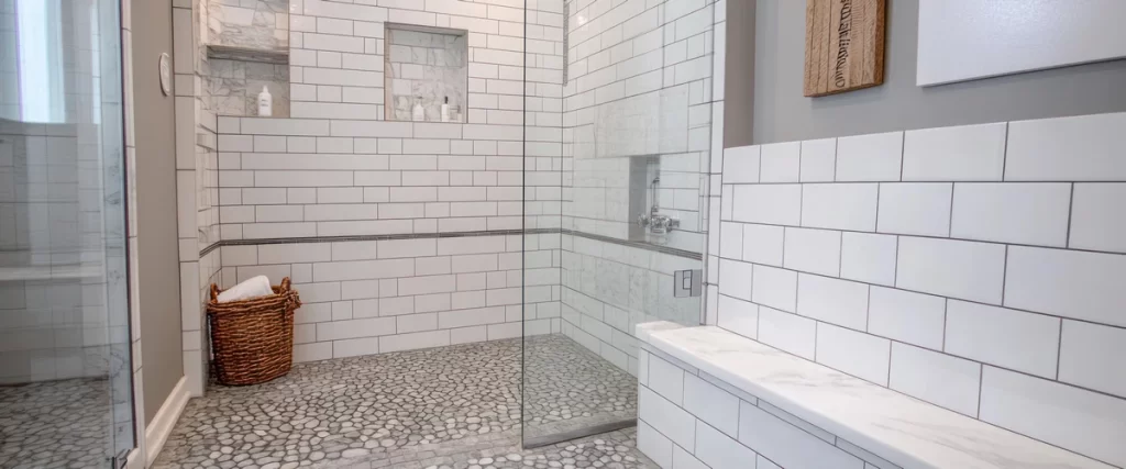 A bright bathroom featuring white subway tile walls, a glass enclosure, a built-in bench, and a grey pebble shower floor with a woven basket inside.