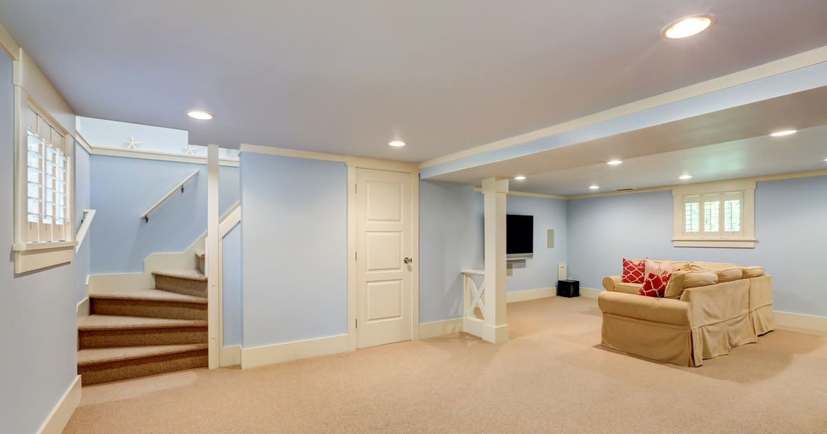 A brightly lit, finished light blue basement featuring beige carpet, a tan sofa with red patterned pillows, a wall-mounted TV, and carpeted stairs, showcasing the quality work of Basement Remodeling Companies metrowest and central massachusetts.