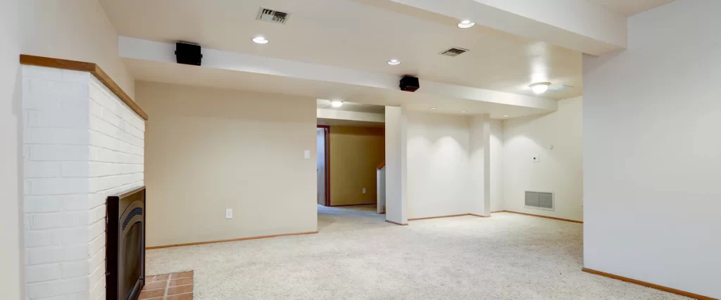 A spacious, empty finished basement ready for furniture, featuring neutral beige walls, light plush carpet, recessed lighting, and a white painted brick fireplace.