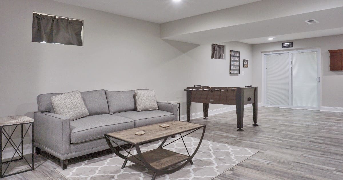 A spacious room showcasing basement remodeling ideas with a comfortable grey sofa, a rustic wood coffee table, and a foosball table near sliding glass doors.