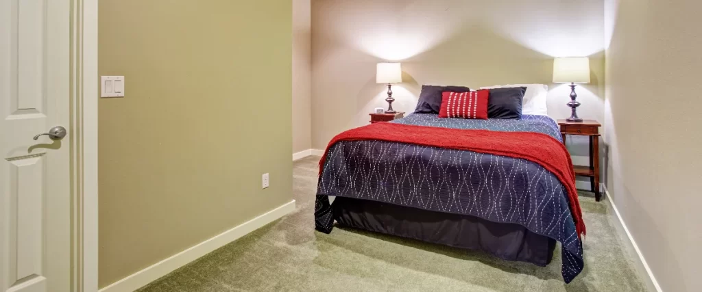 A simple and comfortable basement guest suite bedroom with beige walls, a bed covered in a red and blue patterned blanket, and matching wooden nightstands with lamps.