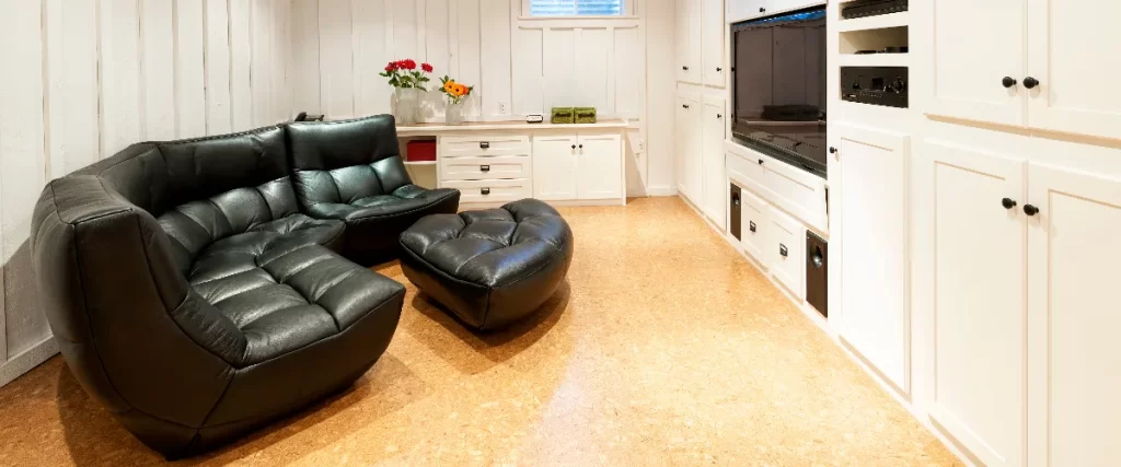 A white paneled basement home theater room featuring custom built-in cabinetry surrounding a TV, cork flooring, and a large black leather modular lounging sofa.