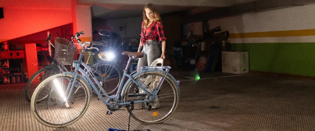 A woman standing next to two bicycles in a dimly lit basement workshop, with the front light blue bicycle's headlight shining brightly in the dark space.