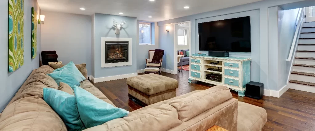 A cozy basement family room featuring a large tan sectional sofa with bright blue pillows, a built-in fireplace, and a large flat-screen TV on a distressed blue console.