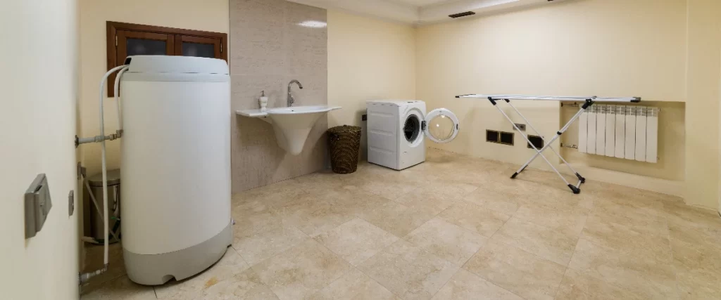A practical basement laundry room with beige tile flooring, featuring a large water heater, a wall-mounted sink, an open washing machine, and a clothing drying rack.