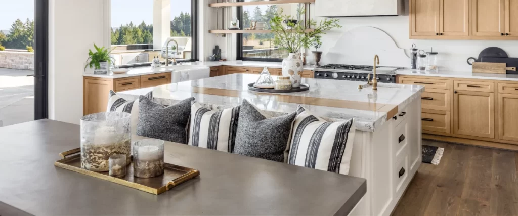 A bright, airy kitchen showcasing a unique custom island with a built-in bench and decorative pillows, set against light wood cabinetry and large scenic windows.