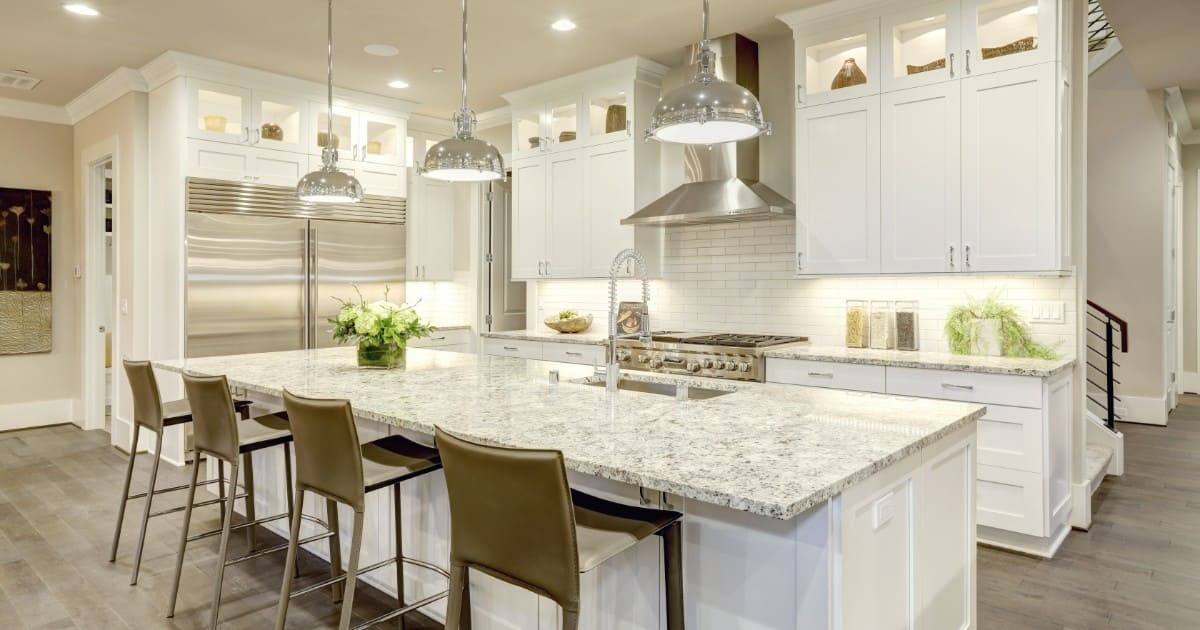 A spacious luxury kitchen featuring white cabinetry, a massive granite island with leather stools, and high-end stainless steel appliances, highlighting factors of kitchen remodel cost.