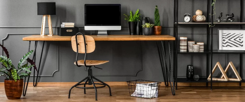 A sleek modern basement home office setup against a dark grey paneled wall, featuring an industrial wooden desk, a swivel chair, a computer monitor, and decorative shelving.