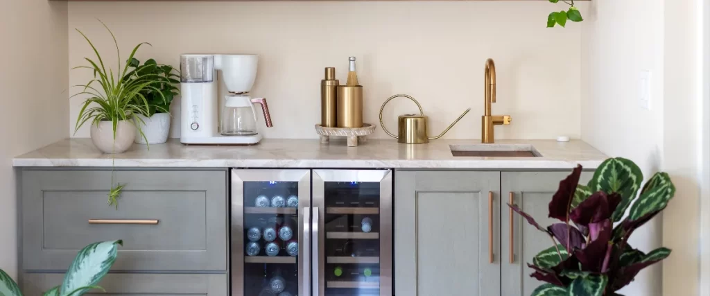 A modern basement wet bar featuring light green cabinetry, a built-in dual-zone beverage cooler, a sink with a gold faucet, and a coffee station.