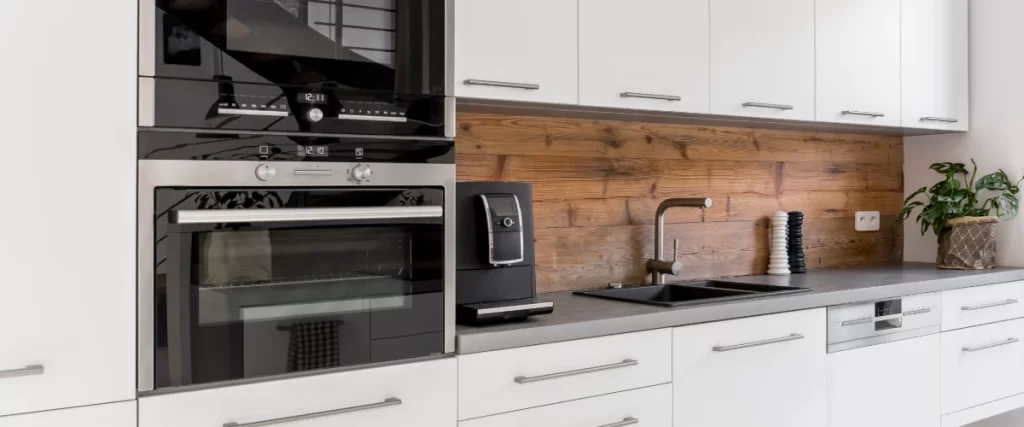 Close-up of a modern kitchen wall featuring built-in stainless steel oven appliances, a black double sink, and a rustic wood plank backsplash.