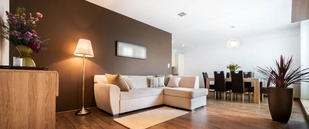 A brightly lit open concept basement rental unit featuring a white modern sofa against a dark brown accent wall, a floor lamp, and a dining table with chairs in the background.