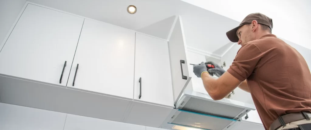 A professional kitchen contractor in a brown uniform using a screwdriver to carefully install or adjust a white upper cabinet door.